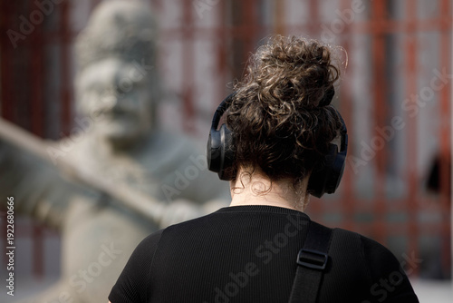 Female tourist with blonde hair tied back and headphones walking, shoulder shot, skin and hair detail, rear view, at Wat Arun.