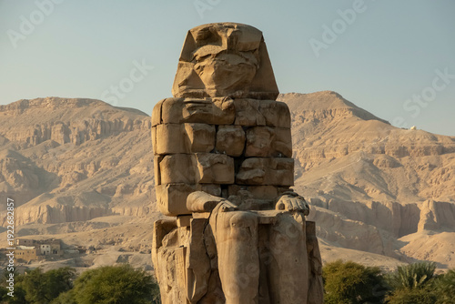 A colossal Egyptian statue of Pharaoh Amenhotep III stands on the site of ancient ruins in Luxor, set in a desert landscape with mountains and a clear sky in the background.