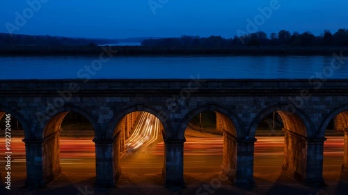Historic stone bridge with traffic light trails at dusk by serene water - 4k video