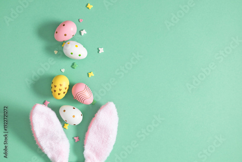 Top view of pink fluffy bunny ears with decorated pastel Easter eggs and candy sprinkles on turquoise background, creative spring holiday flat lay with copy space, festive greeting card template.