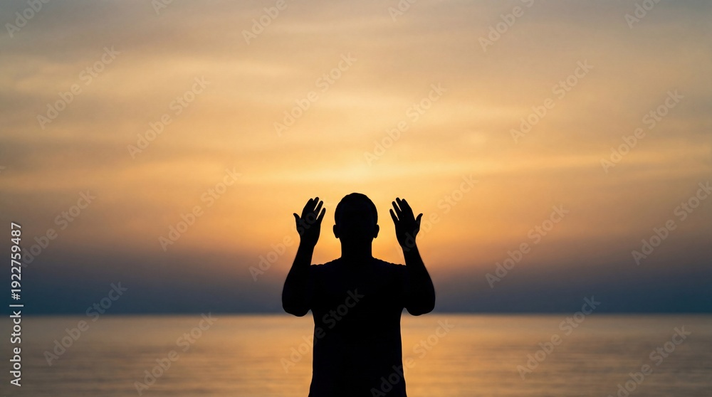 Fototapeta premium Silhouette of a man praying with hands raised towards the sunset over the ocean.