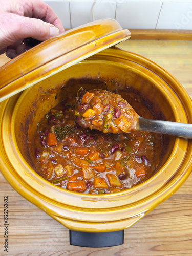Closeup of a hand holding the lid of a slow cooker pot, while lifting out some hot and spicy mixed vegetable chili stew with a large serving spoon.