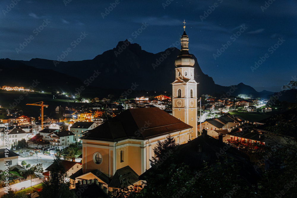 Obraz premium Illuminated Parish Church of Castelrotto at Night – Traditional Alpine Village Architecture with Steeple under Dark Blue Sky in South Tyrol