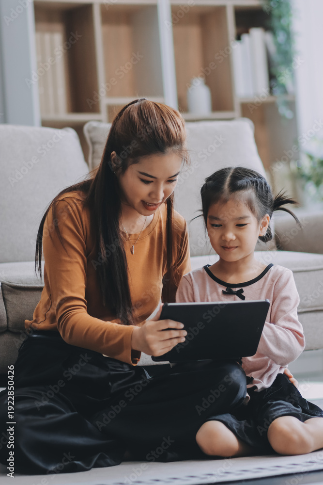 Fototapeta premium A cheerful Asian mother helps her young daughter do homework and color a book at home, while the father works on a laptop in the background. A cozy, bright, and modern family lifestyle.
