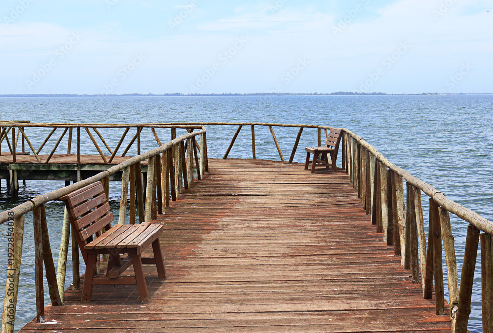 Fototapeta premium IGUABA GRANDE, RIO DE JANEIRO, BRAZIL – January 18, 2025: A rustic wooden deck extends over the tranquil waters of the Araruama Lagoon in Iguaba Grande, Rio de Janeiro, where two benchs face the calm 