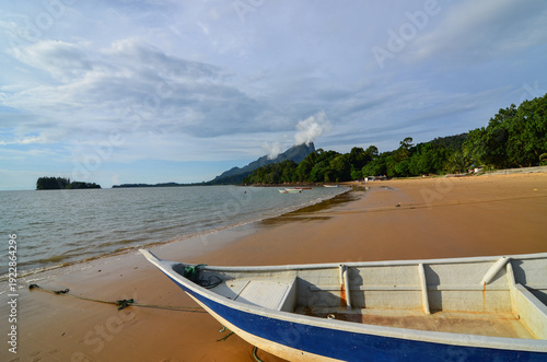 Foreground focus on a blue and white wooden boat resting on a sandy beach with a vast ocean view and mountain scenery in the background.