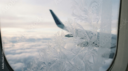 Frosted airplane window with ice feather patterns showing clouds and fuselage edge outside on white soft even light creating calm contemplative mood.