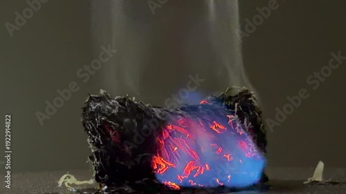 A small bundle of fiber smolders with deep red embers and a vivid blue flame. The glowing heat creates a sense of raw energy and quiet intensity. It is a mesmerizing and powerful sight.