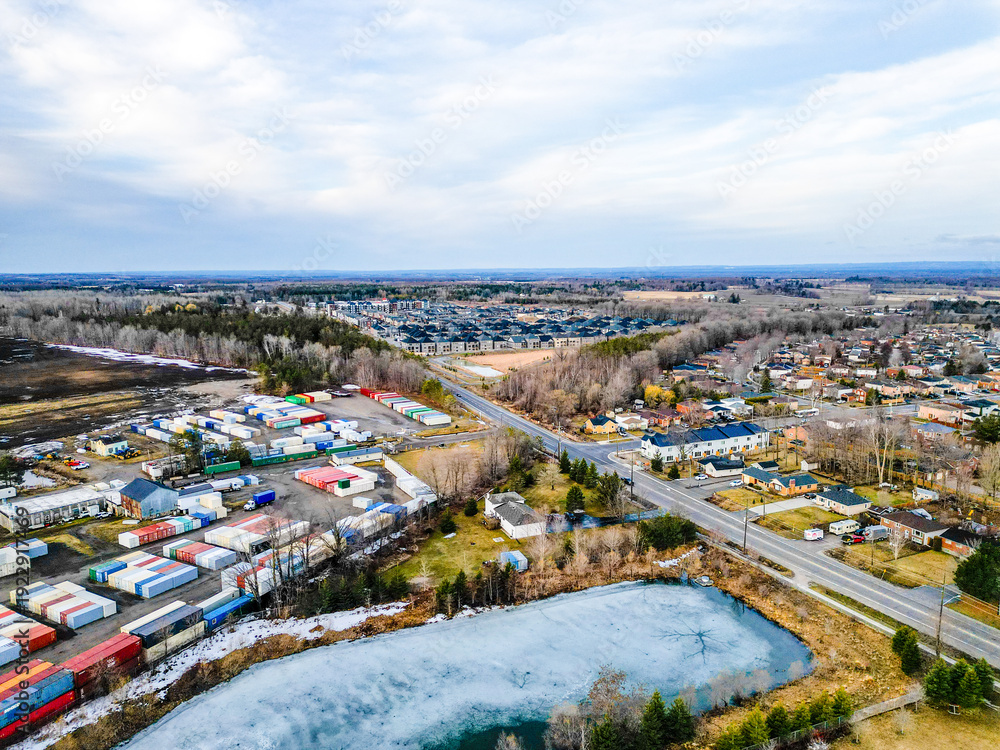 Fototapeta premium Industrial storage yard with rows of shipping containers by a frozen pond and road, bordering houses and new apartment complexes in barrie, ontario, showing urban growth and infrastructure