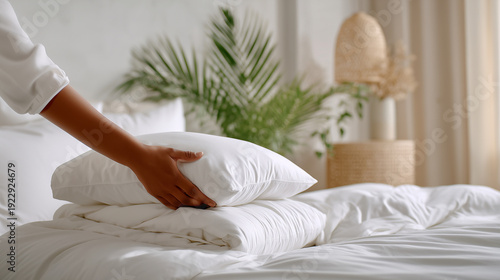 Person arranging fresh white pillow on clean bed with soft white bedding in bright minimalist bedroom with green plant, perfect hotel housekeeping and home comfort concept.