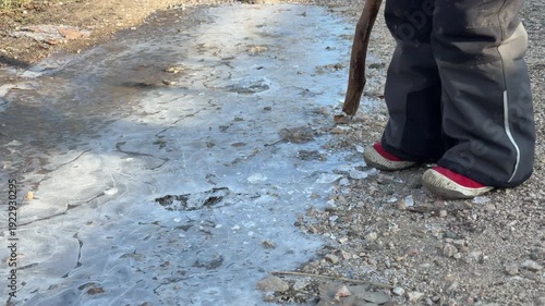 A child firmly holds a wooden branch to break through a layer of frozen ice on a dirt path. This quiet outdoor moment captures the simple joy and focused curiosity of a cold winter morning.