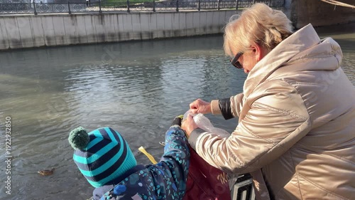 An adult woman and a young child in winter coats reach into a bag together. They are feeding several ducks swimming in a city canal. The sun shines on the water and their warm clothing.