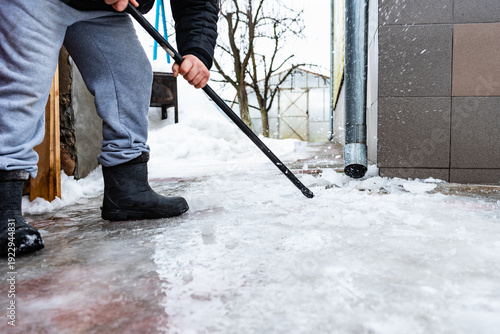 Fotografia Worker on roof with crowbar breaking and removing ice covered sidewalk formed by melting water from drain pipe during winter thaw