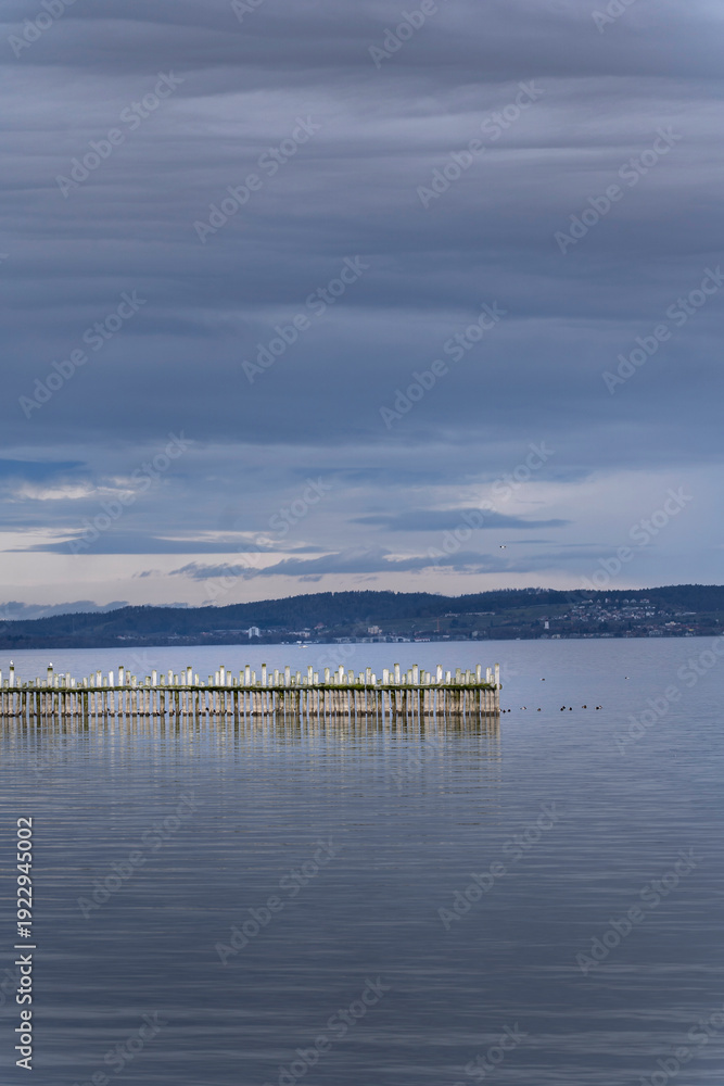 Obraz premium Minimalist wooden pier piles in calm water of Lake Constance. Moody overcast sky, blue grey tones, peaceful landscape. Perfect for nature background, travel editorial or tranquil zen atmosphere