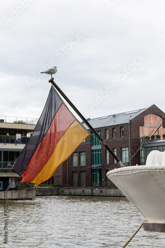 LEER, GERMANY - SEPTEMBER 11, 2025: A seagull perched on a flagpole with the German national flag on the stern of a boat in the harbor, with city buildings and water in the background.