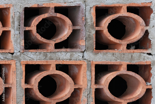 A close-up of a terracotta cobogo wall, showing a geometric pattern of circular and rectangular openings. Rough reddish-brown clay blocks and grey mortar. 