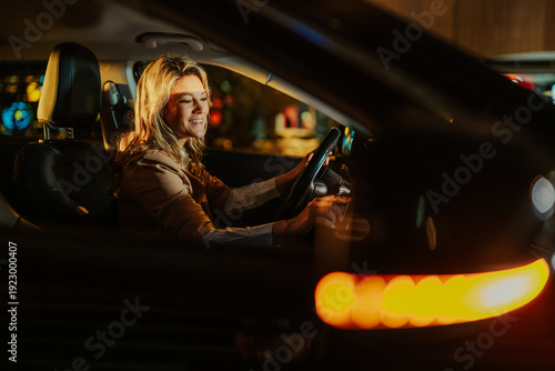 Obraz na plátne Smiling woman driving car at night in city, enjoying the ride with urban lights