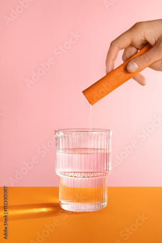 A hand dissolving an orange stick powder in water on pink background.