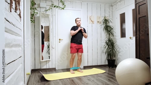 A man doing side lunges with a rubber band on his feet while standing on a yoga mat at home. Fitness routine. Healthy lifestyle, home fitness, daily sport practicing concept. High quality 4k footage.