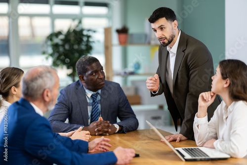 Man stands in front of business colleagues, tells report, proves vision of situation, defends way to solve problems. People are sitting at table, and participant is standing up and took floor. .