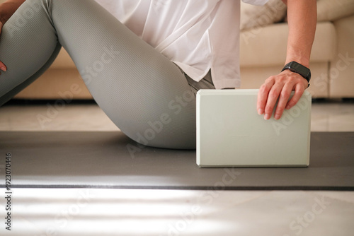 Middle aged woman using yoga block during home workout on mat. Healthy lifestyle concept, fitness stretching, and pilates exercise at home.