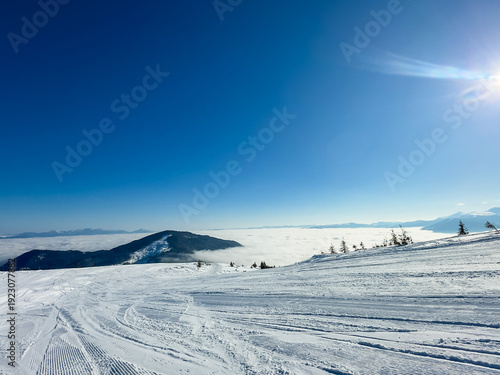 Snow-covered mountain slopes under clear blue sky with clouds below and bright sunlight shining above the landscape