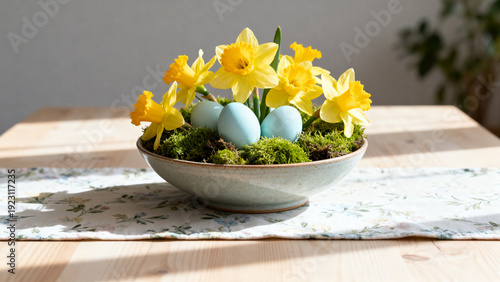 Pale blue bowl of bright daffodils and moss with three light-blue eggs, sunlit spring centerpiece for seasonal presentation