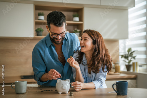 Obraz na plátně Happy couple saving money in piggy bank at home kitchen