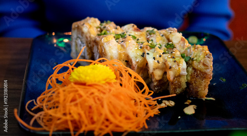 Sushi appetizer dish filled with beef and chicken with dressing, topped with fried bacon and carrot threads with chipotle sauce on a black plate