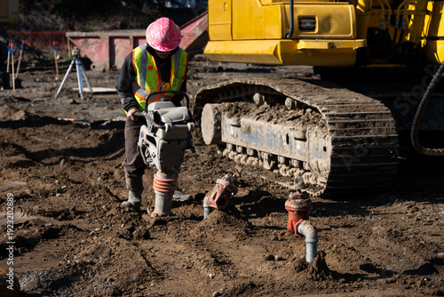 Construction worker using a compactor rammer machine to manually pack the fresh dirt covering trench a newly installed water pipeline, new municipal water distribution utility project work
