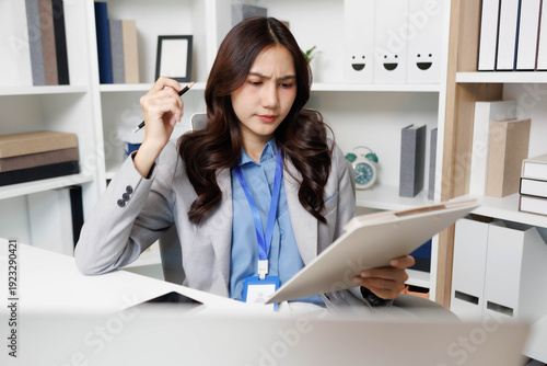 A woman is sitting at a desk with a stack of papers in front of her. She is looking at a piece of paper and she is in a state of confusion or frustration
