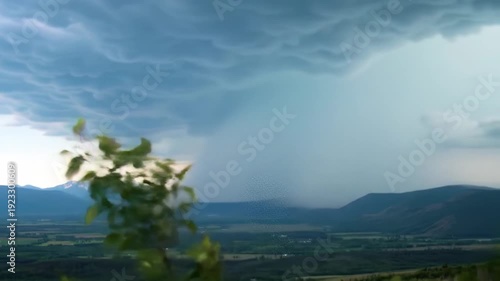 Dramatic storm clouds gather over a lush valley, casting shadows on the distant mountains and fields below.
