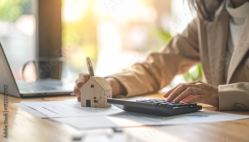 Woman calculating home loan or mortgage payments with a calculator and a small house model on a wooden desk.