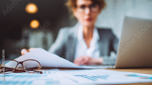A woman is sitting at a desk with a laptop and a piece of paper. She is wearing glasses and she is focused on the paper