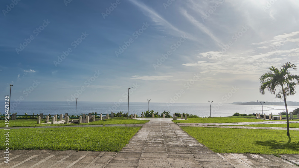 Obraz premium Park by the ocean. Paved footpaths, green grass on lawns. An ancient cannon is visible in the distance, surrounded by a fence. The barrel is aimed at the sea. Cuba. Havana. Battery of Santa Clara 