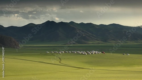 Mongolian ger village spreads across open steppe grassland beneath dark mountain ridge. Crowded yurt settlement lines the green plain, forming a nomad campsite near distant peaks.