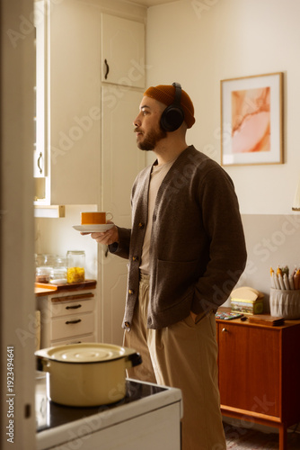 A bearded man in a brown cardigan and orange beanie relaxes in a cozy kitchen, wearing headphones and holding a cup of coffee. Warm, aesthetic lifestyle photography.