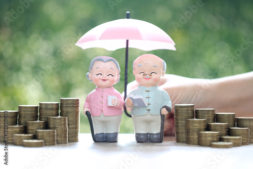Senior couple figures under pink umbrella with stacks of coins on green background. Concept for retirement insurance, financial protection, elderly wealth management and pension security.
