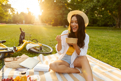 Woman sitting on blanket, smiling as eating fresh salad from bowl during picnic in sunny summer park, fruits and open book nearby on plaid
