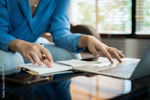 A woman is writing in a notebook and laptop. She is wearing a blue shirt and has a pen in her hand. The scene suggests that she is working on a project or writing something important
