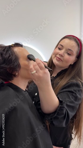 Professional makeup artist applying eye makeup cosmetics to female senior client during beauty treatment session in salon demonstrating MUA work technique for glamour makeover service preparation