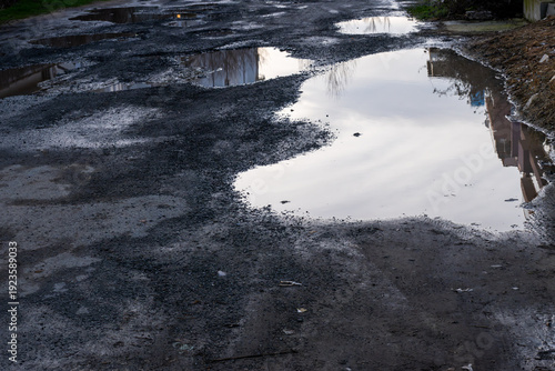 a heavily damaged and uneven dirt road riddled with large, muddy potholes filled with rainwater, reflecting the bright sky and nearby structures on the water's surface.