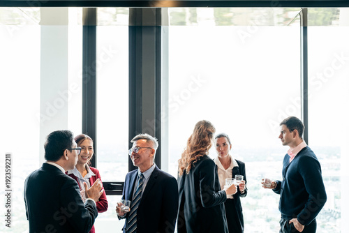 Slika na platnu Professionals networking by large windows in a high-rise office, chatting and holding drinks with a bright city view in the background
