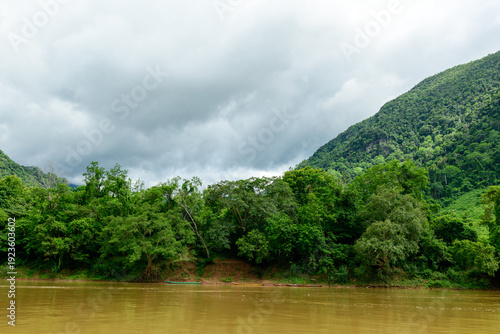 Heavy gray clouds gather above the muddy Nam Ou River and dense green forest near Muang Ngoi in northern Laos. Lush hills rise in the background, creating a dramatic landscape with vibrant vegetation