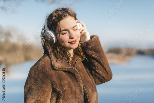 Young woman listening to music with headphones by the sea on a sunny day. Relaxed mood, closed eyes, enjoying sound and nature, modern lifestyle and calm outdoor moment.