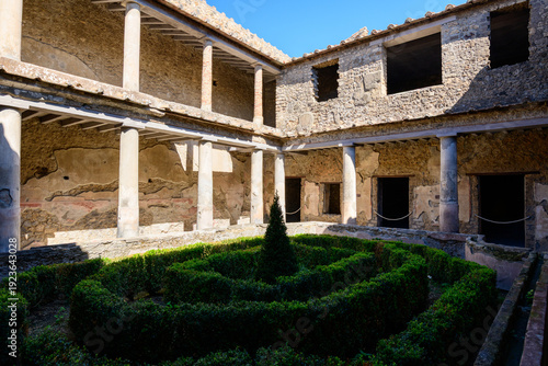 Canvas-taulu A sunlit courtyard with manicured boxwood hedges and a central topiary is framed by ancient stone columns in the House of the Lovers, Pompeii, Italy