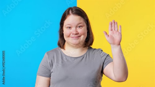 Smiling young woman with Down syndrome waving hand in greeting gesture on a colorful background.