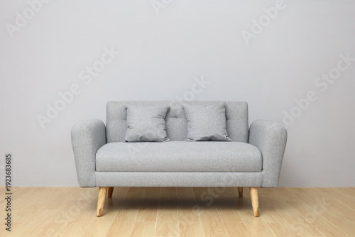 A minimalist light grey fabric two-seater sofa with wooden legs and two matching throw pillows, set against a plain grey wall and light wooden floor