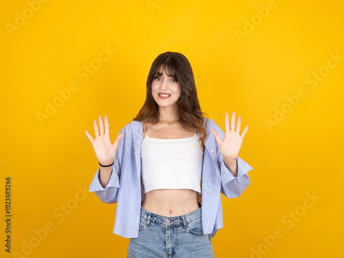 Foto Concerned young woman showing stop gesture with both palms, looking at camera with hesitant expression against yellow background