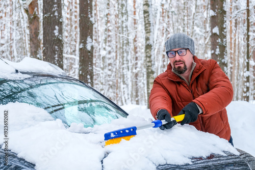 Фотография driver preparing car for trip, man cleaning snow from hood with brush in winter